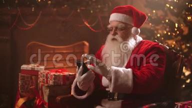 圣诞老人坐在圣诞树背景的椅子上，用智能手机查看他的网上银行